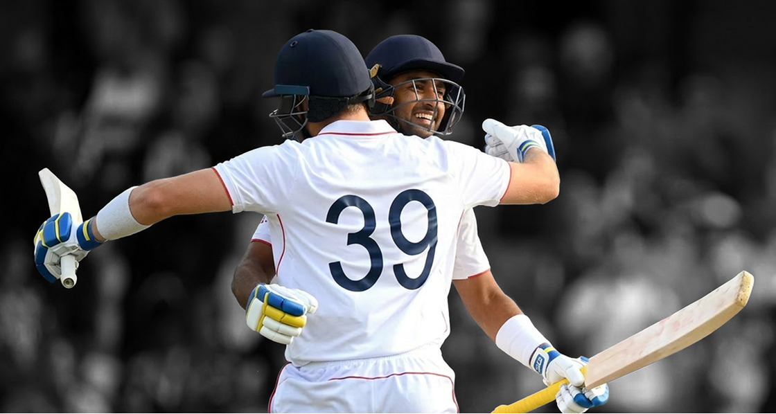 Two pro cricketers in white uniforms hugging in a crowded stadium one holding a bat